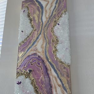 Może przedstawiać: Abstrakcyjna grafika ścienna z motywem geody w odcieniach fioletu, złota i bieli. Grafika ma teksturowaną powierzchnię z kryształowymi akcentami i złotymi detalami brokatu. Element jest zamontowany na drewnianej ramie.