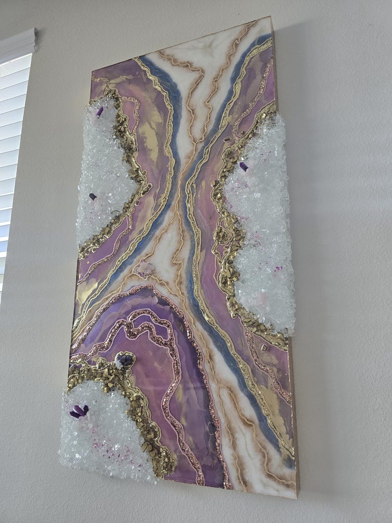 Może przedstawiać: Abstrakcyjna grafika ścienna z motywem geody w odcieniach fioletu, złota i bieli. Grafika ma teksturowaną powierzchnię z kryształowymi akcentami i złotymi detalami brokatu. Element jest zamontowany na drewnianej ramie.