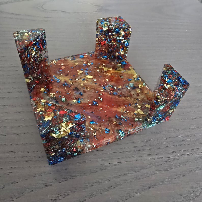 Pode incluir: Um suporte de telefone decorativo em resina com um design &uacute;nico. O suporte apresenta uma mistura de flocos vermelhos, castanhos, azuis e dourados embutidos na resina transparente. Possui uma base triangular com dois suportes verticais.