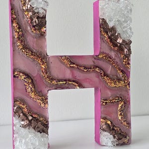 May include: A decorative letter "H" in a vibrant pink hue, adorned with a layered design of pink, gold glitter, and brown accents. The edges are embellished with clear and brown crystal-like pieces, creating a unique, textured appearance.