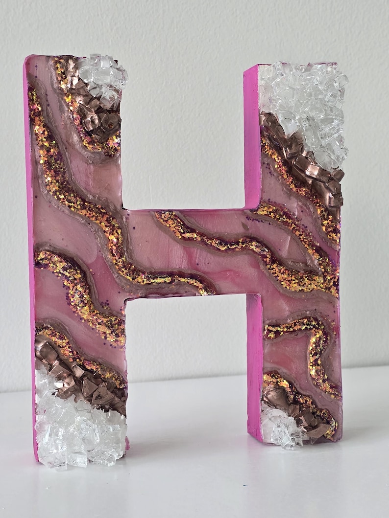 May include: A decorative letter "H" in a vibrant pink hue, adorned with a layered design of pink, gold glitter, and brown accents. The edges are embellished with clear and brown crystal-like pieces, creating a unique, textured appearance.
