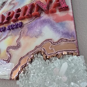 Puede incluir: Una pieza de arte decorativa de resina con el nombre "Selina" en letras en relieve cubiertas de purpurina. La obra de arte presenta un dise&ntilde;o colorido y veteado con tonos rosas, morados y dorados, y est&aacute; adornada con cristales transparentes.