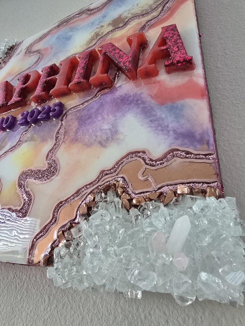 Puede incluir: Una pieza de arte decorativa de resina con el nombre "Selina" en letras en relieve cubiertas de purpurina. La obra de arte presenta un dise&ntilde;o colorido y veteado con tonos rosas, morados y dorados, y est&aacute; adornada con cristales transparentes.