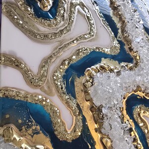 Op de afbeelding: Abstract kunstwerk met een geode-achtig ontwerp. Het kunstwerk heeft lagen van witte, teal en marineblauwe hars, geaccentueerd met gouden glitter en bladgoud. Gebroken glas voegt textuur toe.