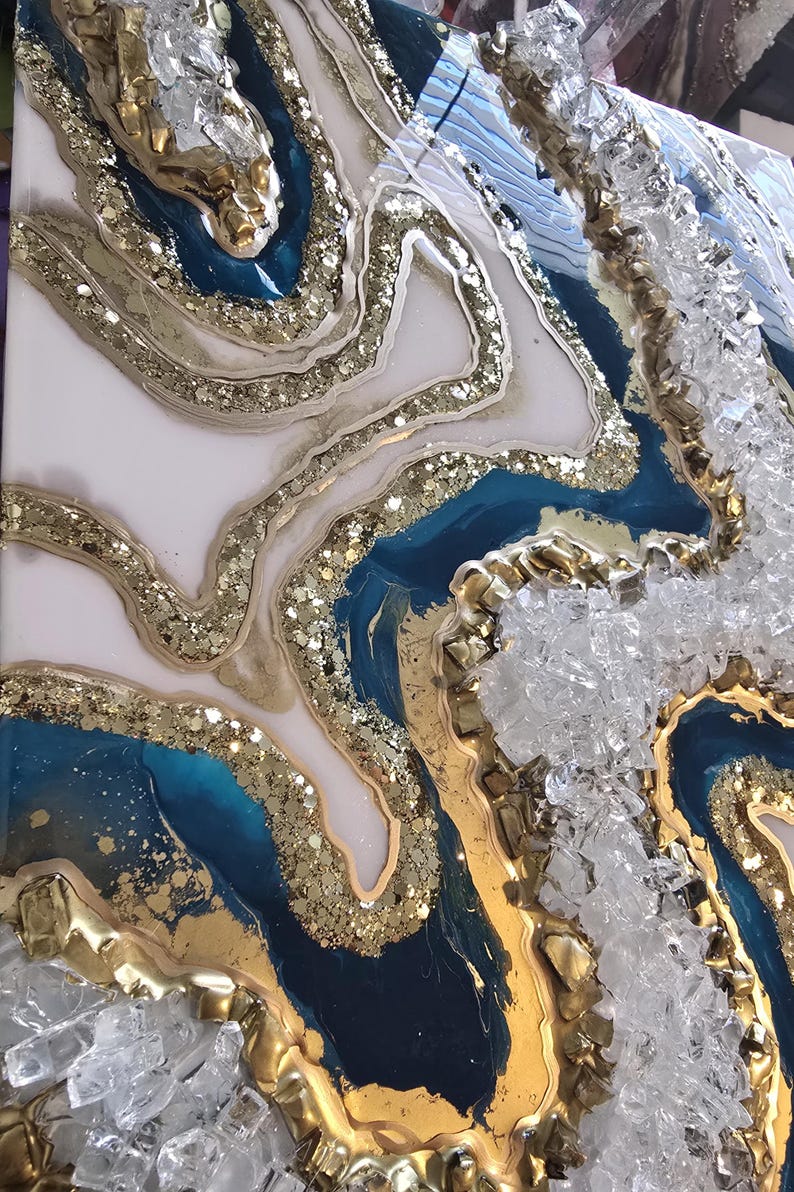 Op de afbeelding: Abstract kunstwerk met een geode-achtig ontwerp. Het kunstwerk heeft lagen van witte, teal en marineblauwe hars, geaccentueerd met gouden glitter en bladgoud. Gebroken glas voegt textuur toe.