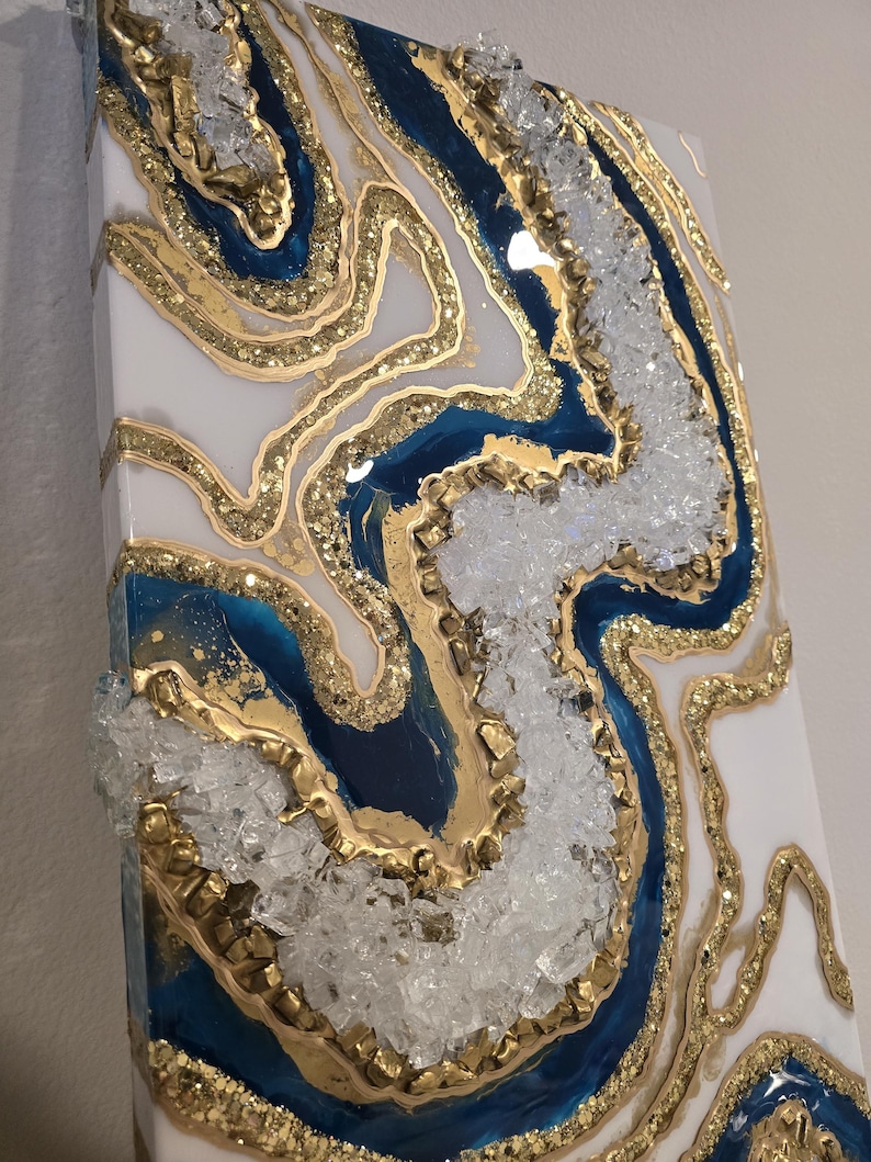 Op de afbeelding: Abstracte wandkunst met een geode-ontwerp in wit, marineblauw en goud. Het getextureerde kunstwerk bevat kristallen en glitter, waardoor een luxueus en opvallend stuk voor interieurdecoratie ontstaat.