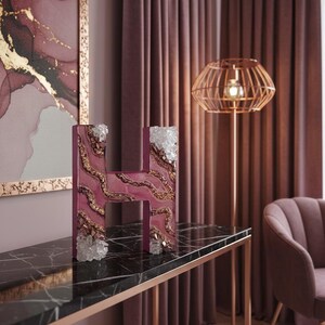 May include: A decorative letter "H" in a deep magenta hue, adorned with gold accents and clear crystal embellishments. The letter sits on a black marble table with a rose gold frame, complementing the room's decor.