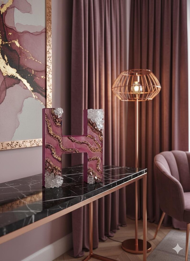 May include: A decorative letter "H" in a deep magenta hue, adorned with gold accents and clear crystal embellishments. The letter sits on a black marble table with a rose gold frame, complementing the room's decor.