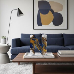 Puede incluir: Una letra decorativa "N" hecha de resina, con remolinos azules y dorados y detalles de cristal. La letra se exhibe en una bandeja de m&aacute;rmol sobre una mesa de centro de madera en un sal&oacute;n.
