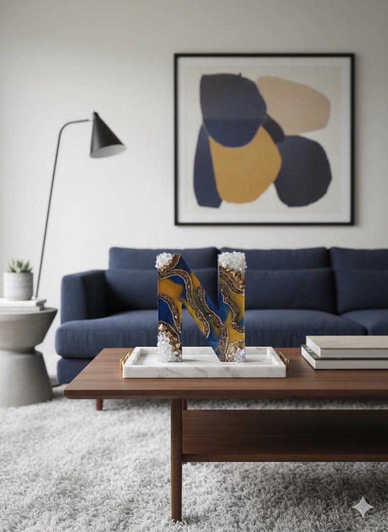 Puede incluir: Una letra decorativa "N" hecha de resina, con remolinos azules y dorados y detalles de cristal. La letra se exhibe en una bandeja de m&aacute;rmol sobre una mesa de centro de madera en un sal&oacute;n.