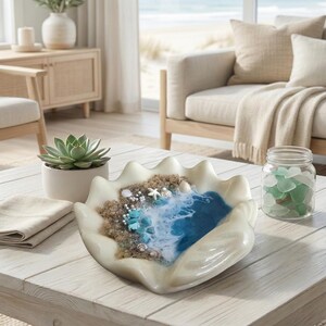 May include: A decorative seashell-shaped bowl with a beach scene, including sand, seashells, and a blue resin ocean. A succulent in a white pot, a folded beige napkin, and a jar of sea glass are on the white wooden table.