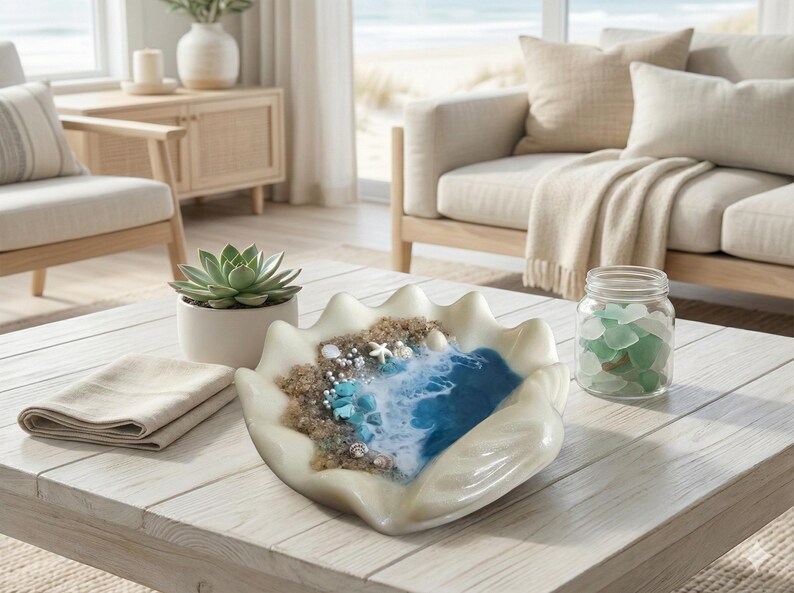 May include: A decorative seashell-shaped bowl with a beach scene, including sand, seashells, and a blue resin ocean. A succulent in a white pot, a folded beige napkin, and a jar of sea glass are on the white wooden table.