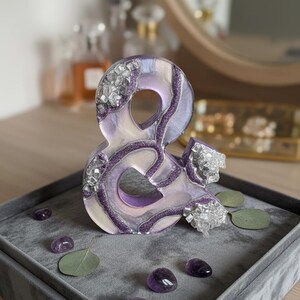 May include: A decorative ampersand sculpture in shades of purple, white, and silver, adorned with crystals and glitter. The sculpture sits on a gray velvet tray with amethyst stones and eucalyptus leaves.