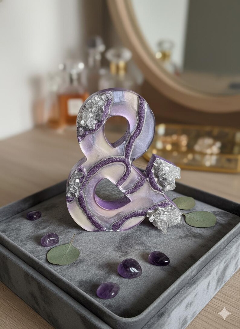 May include: A decorative ampersand sculpture in shades of purple, white, and silver, adorned with crystals and glitter. The sculpture sits on a gray velvet tray with amethyst stones and eucalyptus leaves.