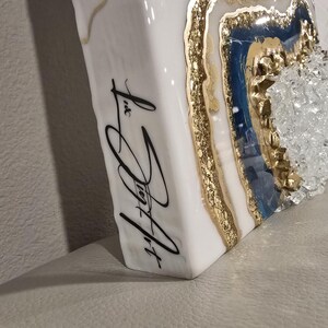 Op de afbeelding: Wit, rechthoekig kunstwerk met een geode-ontwerp. De geode heeft gouden, blauwe en heldere kristalelementen. De zijkant van het stuk heeft zwarte cursieve tekst met de tekst "Liz Joy Art".