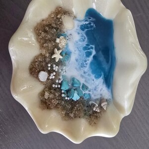 May include: A decorative seashell-shaped dish with a beach scene. The dish features a sandy area with small shells, a starfish, and turquoise stones. Blue and white resin represents the ocean. The dish is pearlescent white.