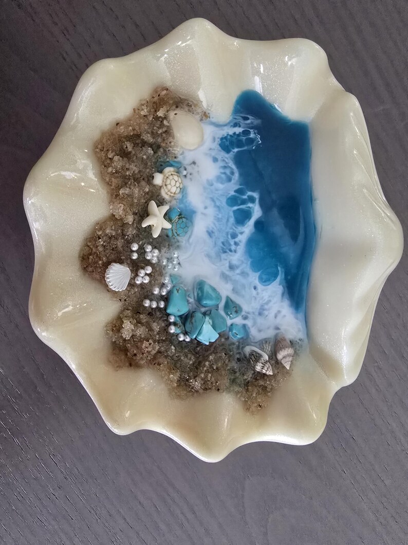 May include: A decorative seashell-shaped dish with a beach scene. The dish features a sandy area with small shells, a starfish, and turquoise stones. Blue and white resin represents the ocean. The dish is pearlescent white.