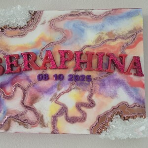 Puede incluir: Lienzo decorativo con un dise&ntilde;o abstracto colorido en tonos rosa, morado y naranja. El nombre "SERAPHINA" se muestra en letras rosas brillantes en relieve, junto con la fecha "08 10 2025". Acentos de cristal transparente adornan las esquinas.