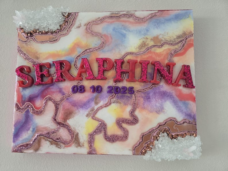 Puede incluir: Lienzo decorativo con un dise&ntilde;o abstracto colorido en tonos rosa, morado y naranja. El nombre "SERAPHINA" se muestra en letras rosas brillantes en relieve, junto con la fecha "08 10 2025". Acentos de cristal transparente adornan las esquinas.