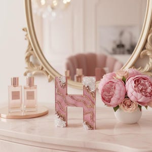 May include: A pink letter "H" with gold glitter accents and crystal embellishments. The letter sits on a pink marble surface, next to a bouquet of pink peonies and two perfume bottles. A gold-framed mirror is in the background.