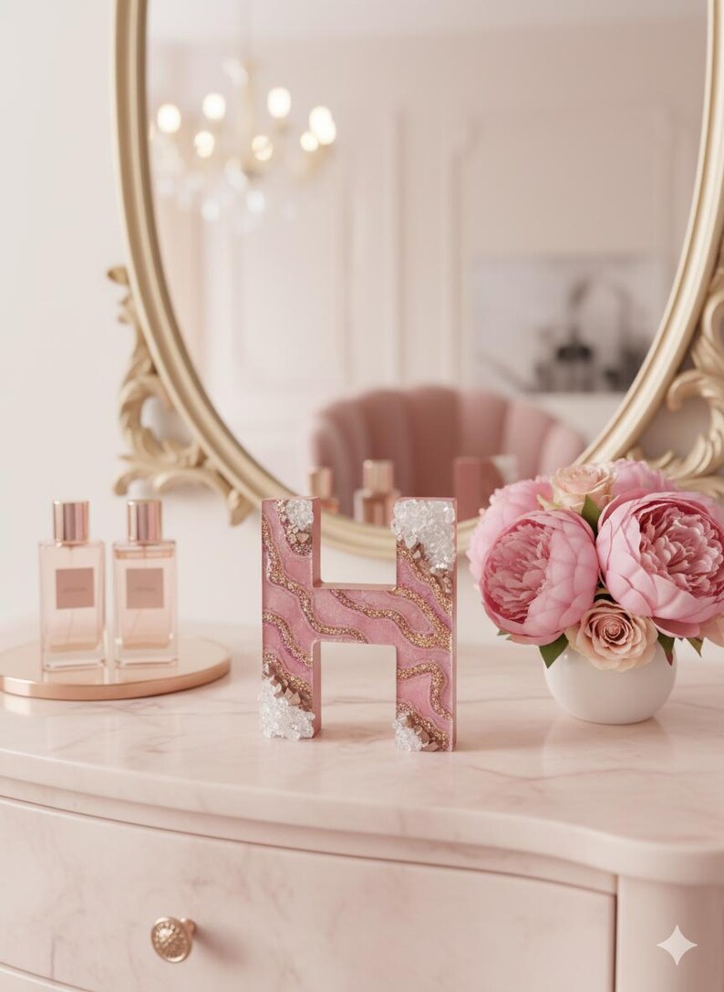 May include: A pink letter "H" with gold glitter accents and crystal embellishments. The letter sits on a pink marble surface, next to a bouquet of pink peonies and two perfume bottles. A gold-framed mirror is in the background.