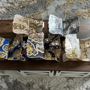 May include: A collection of decorative resin pieces with a unique, abstract design. The pieces feature a variety of colors, including blue, black, white, and gold, with a marbled effect. The pieces are displayed on a dark brown wooden table.