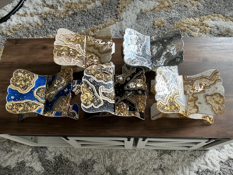 May include: A collection of decorative resin pieces with a unique, abstract design. The pieces feature a variety of colors, including blue, black, white, and gold, with a marbled effect. The pieces are displayed on a dark brown wooden table.