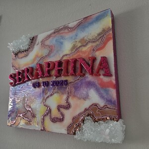 Puede incluir: Una pieza de arte mural cuadrada con un dise&ntilde;o abstracto colorido, con el nombre "SERAPHINA" en letras de purpurina rosa y la fecha "08-10-2023". La obra de arte est&aacute; adornada con detalles de cristal y un borde brillante.