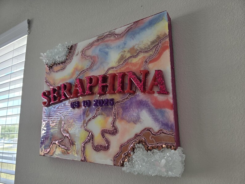 Puede incluir: Una pieza de arte mural cuadrada con un dise&ntilde;o abstracto colorido, con el nombre "SERAPHINA" en letras de purpurina rosa y la fecha "08-10-2023". La obra de arte est&aacute; adornada con detalles de cristal y un borde brillante.