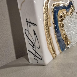 Op de afbeelding: Wit, rechthoekig kunstwerk met een geode-ontwerp. De geode heeft gouden, blauwe en heldere kristalelementen. De zijkant van het stuk heeft zwarte cursieve tekst met de tekst "Liz Joy Art".