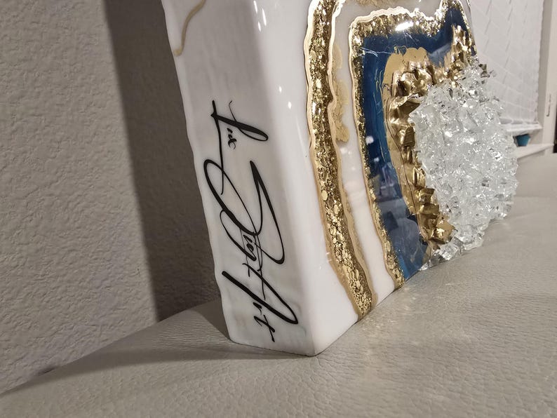 Op de afbeelding: Wit, rechthoekig kunstwerk met een geode-ontwerp. De geode heeft gouden, blauwe en heldere kristalelementen. De zijkant van het stuk heeft zwarte cursieve tekst met de tekst "Liz Joy Art".