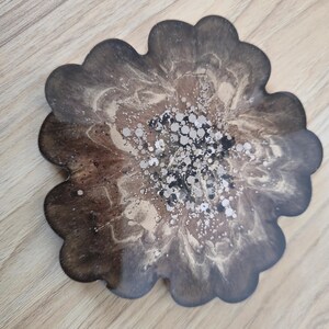 K&ouml;nnte beinhalten: Ein bl&uuml;tenf&ouml;rmiger Untersetzer mit einem dunkelbraunen, beigen und silbernen Design. Der Untersetzer hat einen gewellten Rand und eine gl&auml;nzende Oberfl&auml;che. Die Mitte des Untersetzers ist mit silbernem Glitzer verziert.