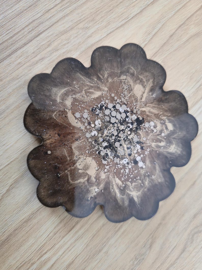 K&ouml;nnte beinhalten: Ein bl&uuml;tenf&ouml;rmiger Untersetzer mit einem dunkelbraunen, beigen und silbernen Design. Der Untersetzer hat einen gewellten Rand und eine gl&auml;nzende Oberfl&auml;che. Die Mitte des Untersetzers ist mit silbernem Glitzer verziert.