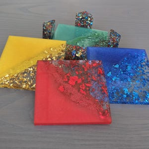 Pode incluir: Um conjunto de bases de copos quadradas coloridas em amarelo, vermelho, azul e verde. Cada base de copo &eacute; incrustada com flocos de folha de ouro ou azul, e algumas t&ecirc;m bordas decorativas correspondentes. As bases de copos est&atilde;o dispostas em uma superf&iacute;cie cinza claro.
