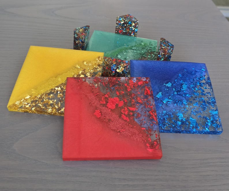 Pode incluir: Um conjunto de bases de copos quadradas coloridas em amarelo, vermelho, azul e verde. Cada base de copo &eacute; incrustada com flocos de folha de ouro ou azul, e algumas t&ecirc;m bordas decorativas correspondentes. As bases de copos est&atilde;o dispostas em uma superf&iacute;cie cinza claro.