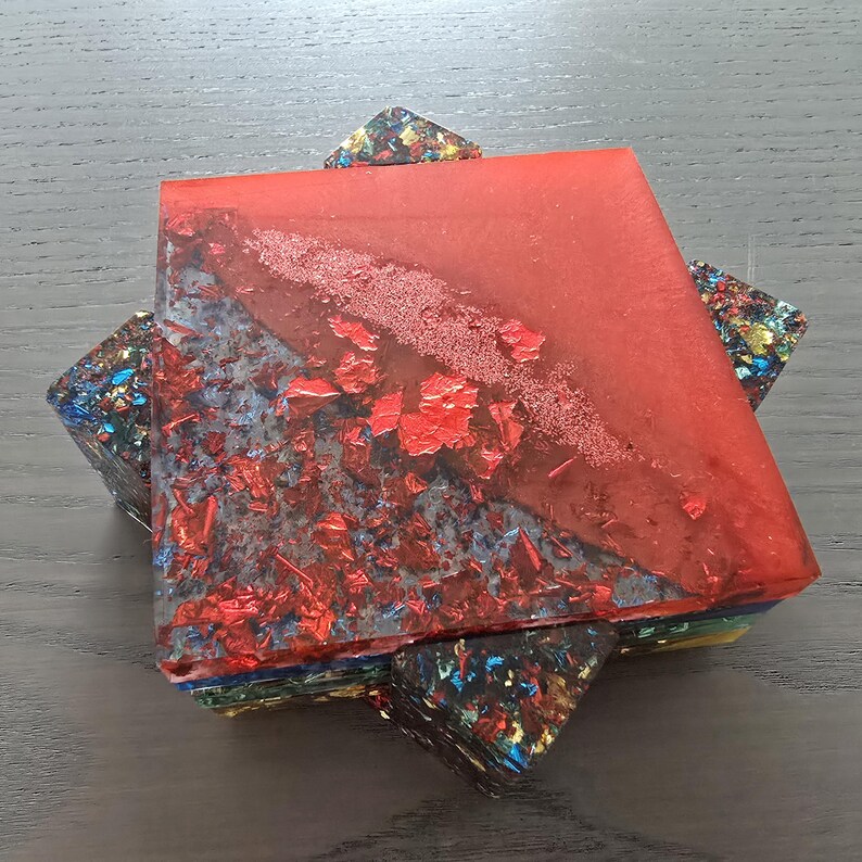 Pode incluir: Uma base para copos geom&eacute;trica com um topo quadrado vermelho incrustado com folha vermelha. A base para copos &eacute; suportada por blocos triangulares coloridos. O design apresenta uma mistura de cores, incluindo vermelho, azul, amarelo e verde, criando um visual moderno e art&iacute;stico.