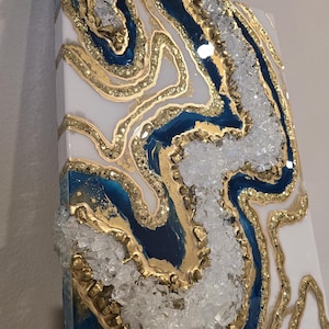 May include: Abstract wall art featuring a geode design. The artwork incorporates white, navy blue, and gold resin, with clear crystal accents. The piece has a textured surface and a glossy finish, creating a luxurious aesthetic.