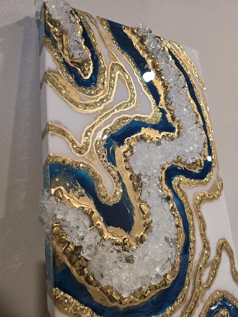 May include: Abstract wall art featuring a geode design. The artwork incorporates white, navy blue, and gold resin, with clear crystal accents. The piece has a textured surface and a glossy finish, creating a luxurious aesthetic.