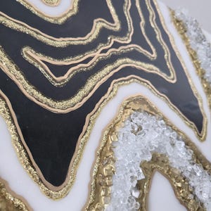 May include: Abstract artwork with a geode design. It features layers of black, white, and gold, with a textured surface. The gold elements include glitter and crystal accents, creating a luxurious aesthetic.