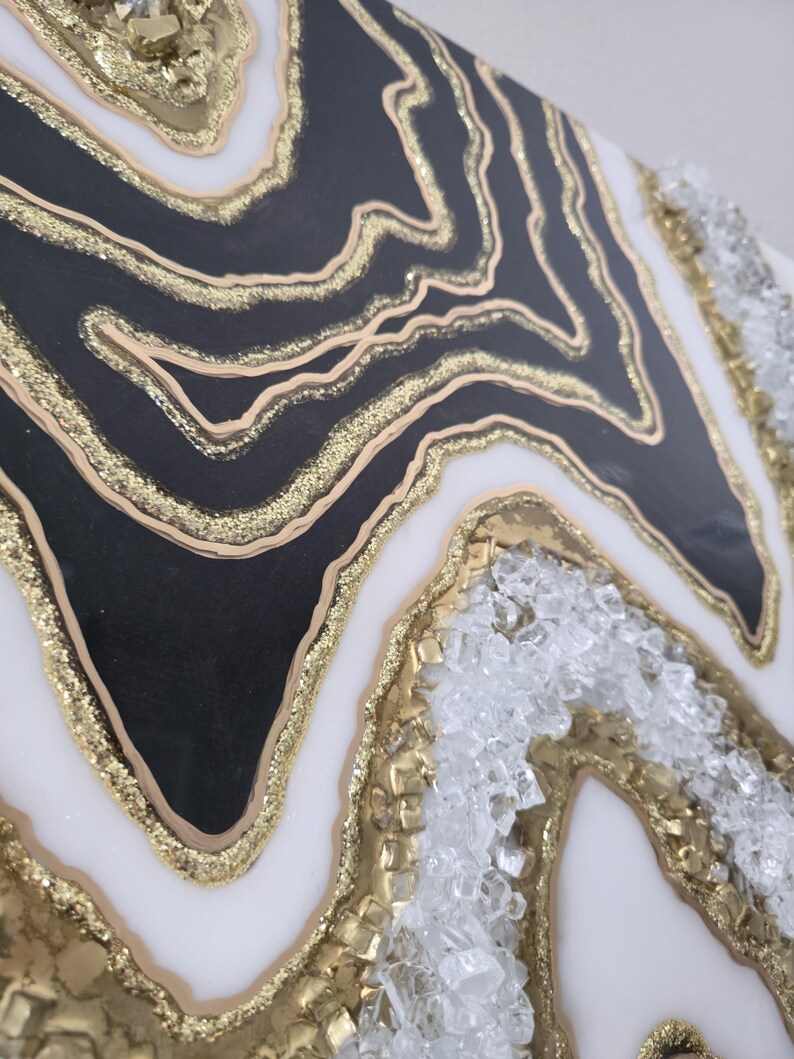 May include: Abstract artwork with a geode design. It features layers of black, white, and gold, with a textured surface. The gold elements include glitter and crystal accents, creating a luxurious aesthetic.