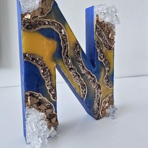 Puede incluir: Una letra decorativa "N" con un exterior azul y un dise&ntilde;o interior amarillo y azul. El interior tiene una apariencia de geoda con detalles dorados y brillantes. Adornos transparentes en forma de cristal en la parte superior e inferior.