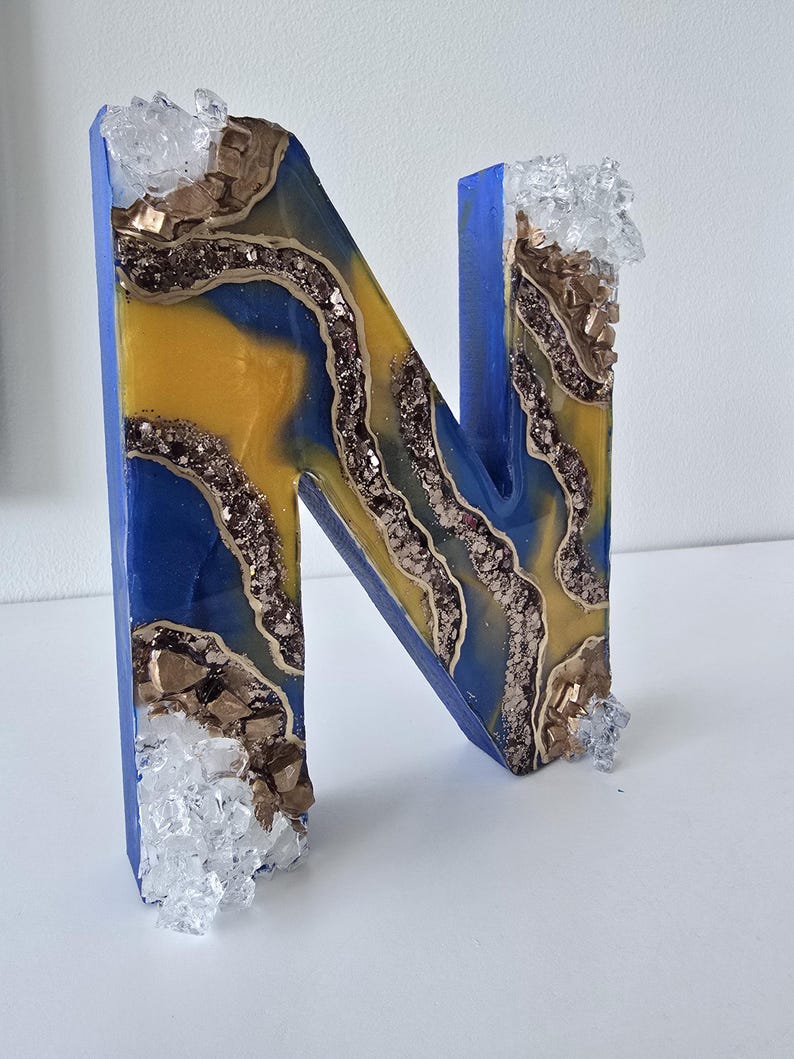Puede incluir: Una letra decorativa "N" con un exterior azul y un dise&ntilde;o interior amarillo y azul. El interior tiene una apariencia de geoda con detalles dorados y brillantes. Adornos transparentes en forma de cristal en la parte superior e inferior.