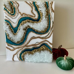 May include: White canvas art with teal, gold, and clear crystal accents. The abstract design features layered resin with a geode-like appearance. Two velvet pumpkins in red and green are next to the artwork.