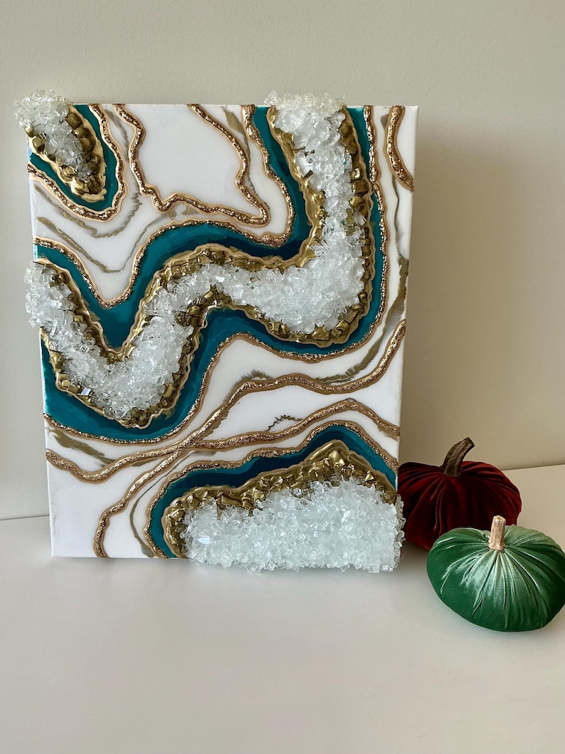 May include: White canvas art with teal, gold, and clear crystal accents. The abstract design features layered resin with a geode-like appearance. Two velvet pumpkins in red and green are next to the artwork.