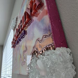 Puede incluir: Una pieza de arte decorativo de pared con un dise&ntilde;o en capas. La obra de arte presenta un fondo colorido con una superficie brillante y texturizada. Letras rojas y brillantes deletrean una palabra. Vidrio triturado y cristales transparentes est&aacute;n adheridos a la obra de arte.