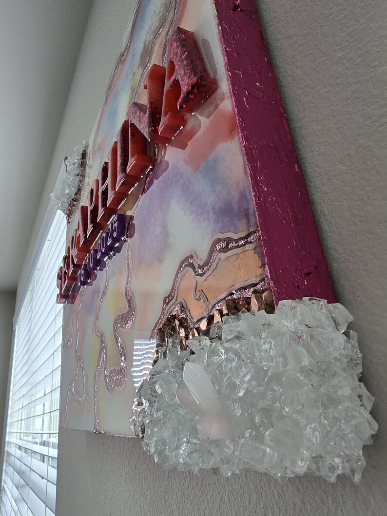 Puede incluir: Una pieza de arte decorativo de pared con un dise&ntilde;o en capas. La obra de arte presenta un fondo colorido con una superficie brillante y texturizada. Letras rojas y brillantes deletrean una palabra. Vidrio triturado y cristales transparentes est&aacute;n adheridos a la obra de arte.