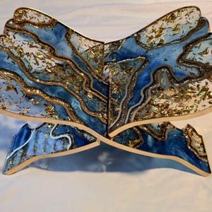 May include: A decorative wooden stand with a blue and white resin design, accented with gold leaf. The stand is shaped like an open book, with a layered, abstract pattern. The edges are outlined in gold.