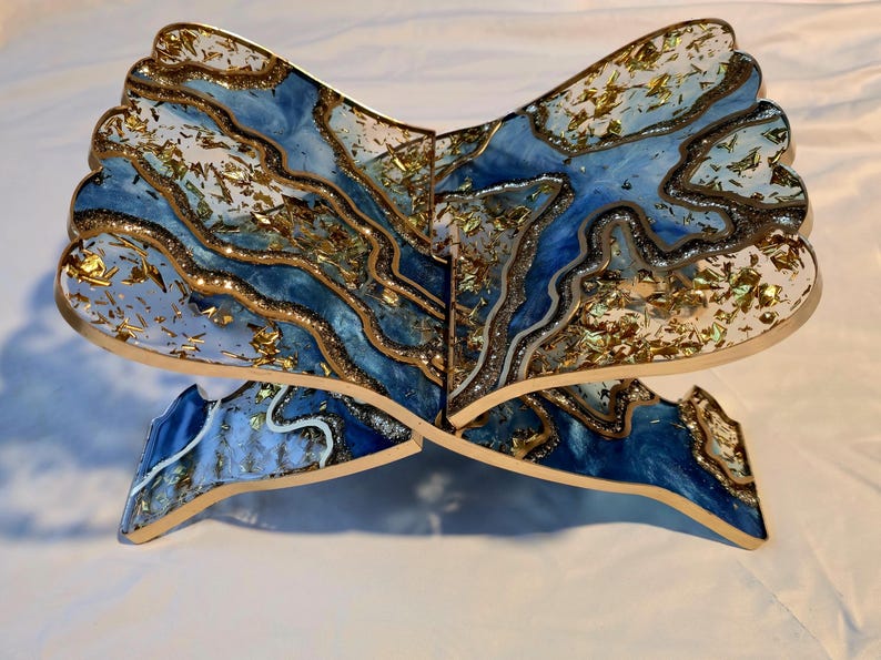 May include: A decorative wooden stand with a blue and white resin design, accented with gold leaf. The stand is shaped like an open book, with a layered, abstract pattern. The edges are outlined in gold.