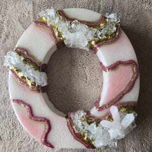 May include: A circular decorative piece with a white base, accented with pink and gold glitter, and clusters of clear crystals. The design features a ring shape with a textured surface, suitable for home decor.