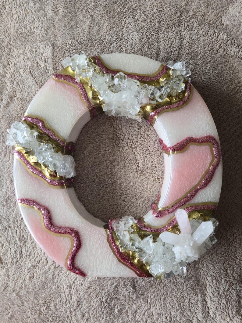 May include: A circular decorative piece with a white base, accented with pink and gold glitter, and clusters of clear crystals. The design features a ring shape with a textured surface, suitable for home decor.